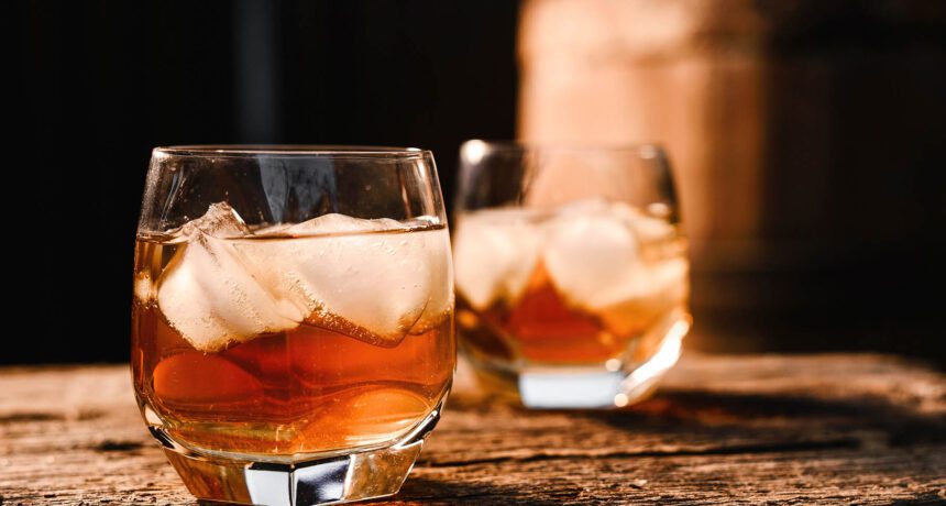 A glass of whiskey with ice on a wooden background close-up in the background of a barrel of whiskey