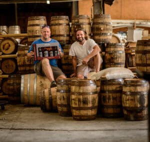 Cam holding a bourbon blending kit next to Aaron sitting on barrels at the og distillery.