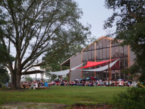 differnet angle back of distillery with old white oak in foreground. large crowd facing the camera watching a comedy show.