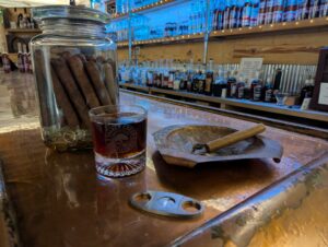 A glass of whiskey sits next to a copper ashtray with a unlit cigar. A large jar of bourbon infused cigars sits behind.